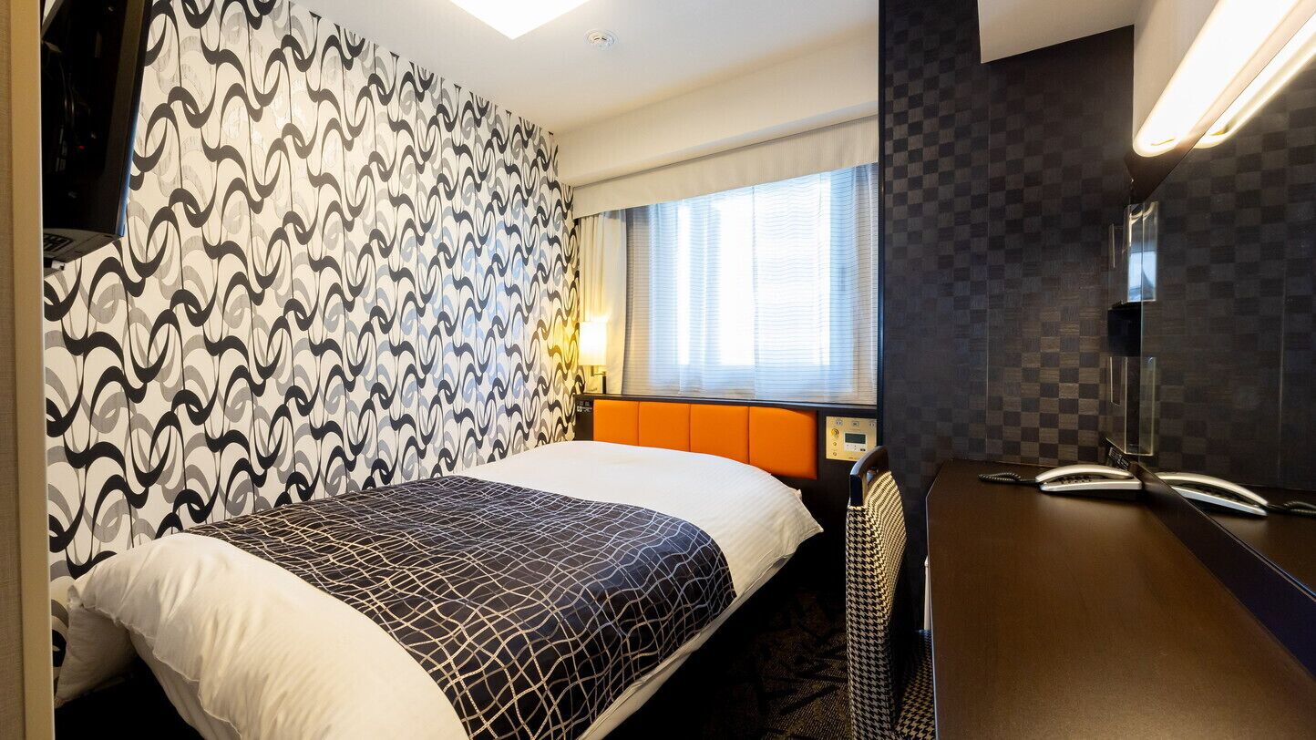 A hotel room with a bed, a desk with a telephone, a window with blinds, and a patterned wall.