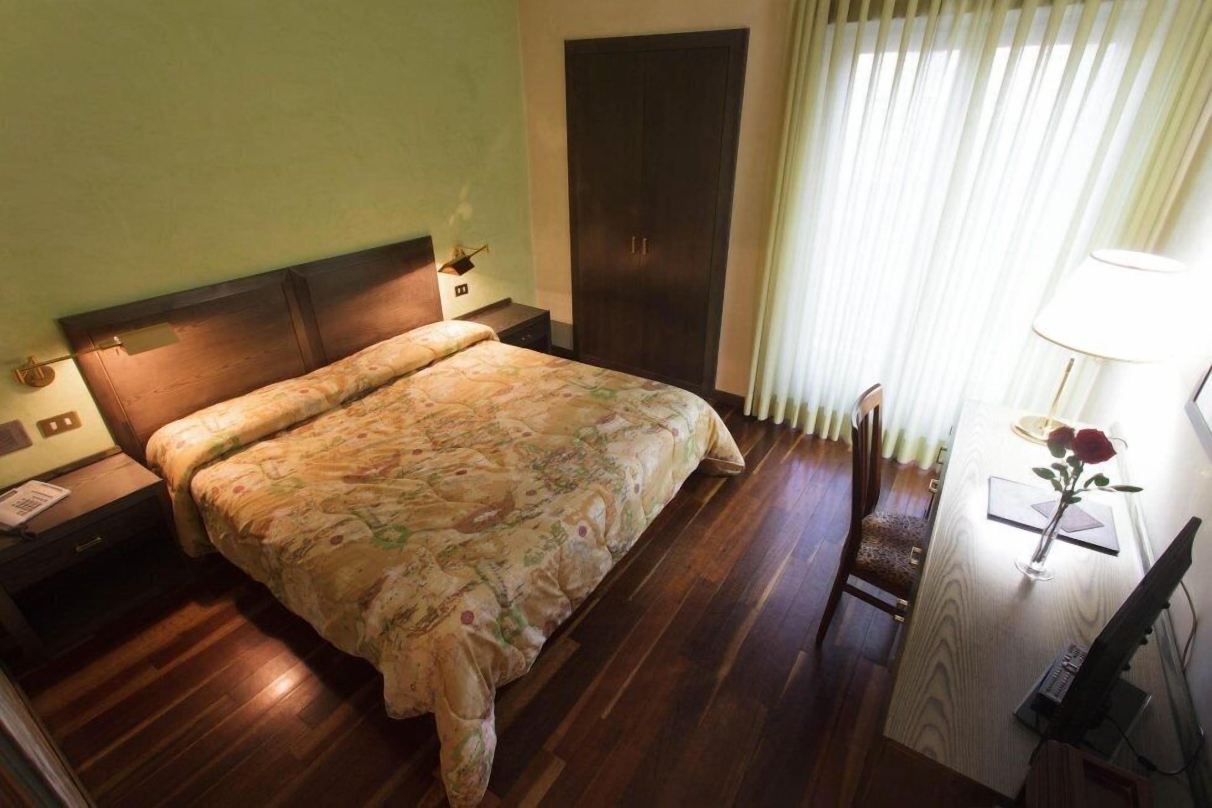 Ein Hotelzimmer mit einem großen Bett, Holzboden, einem Schreibtisch mit Lampe und einem fenster mit Vorhängen.