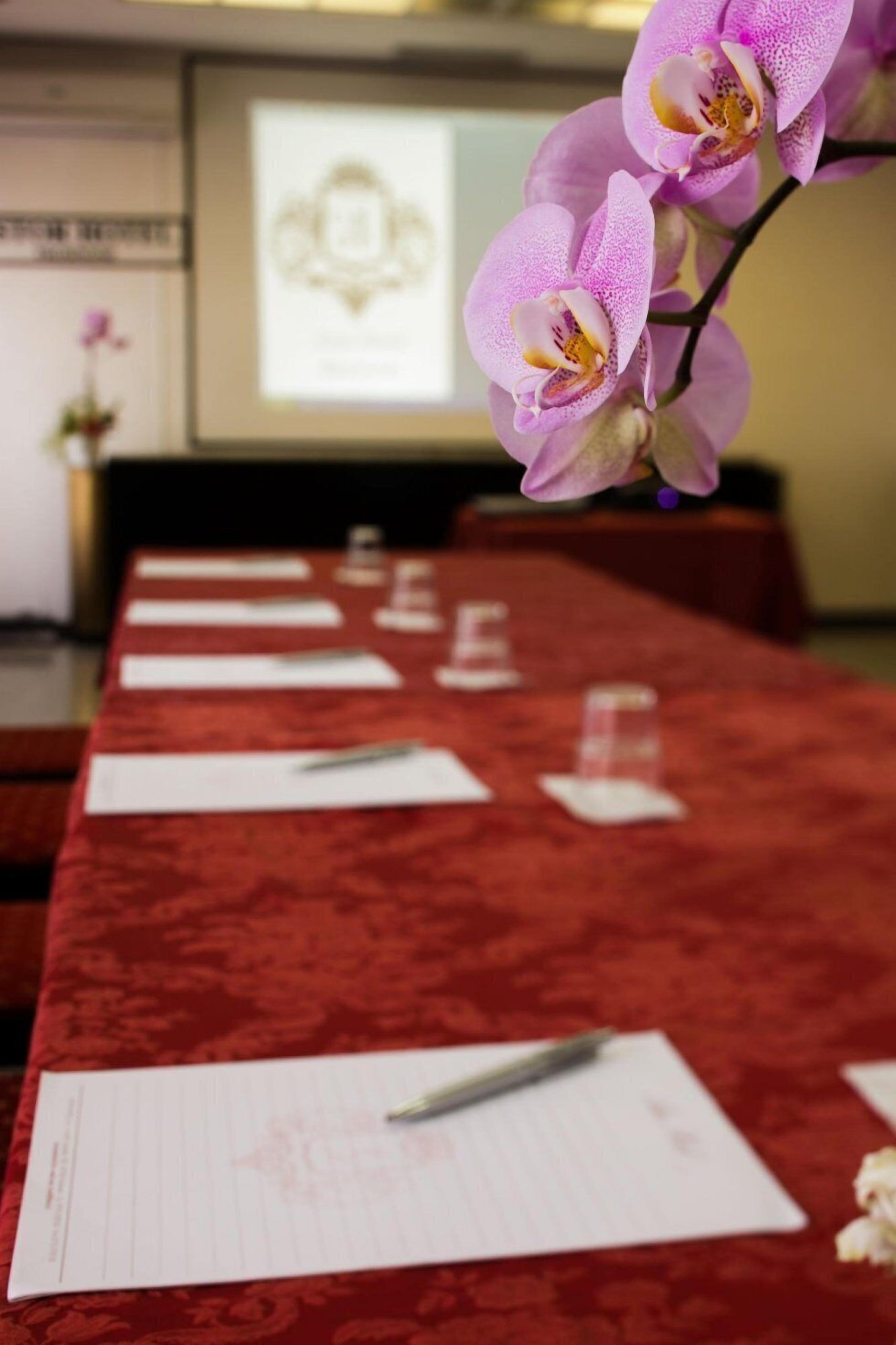 A conference table with documents and pens, a red carpet, and a purple orchid in the foreground.