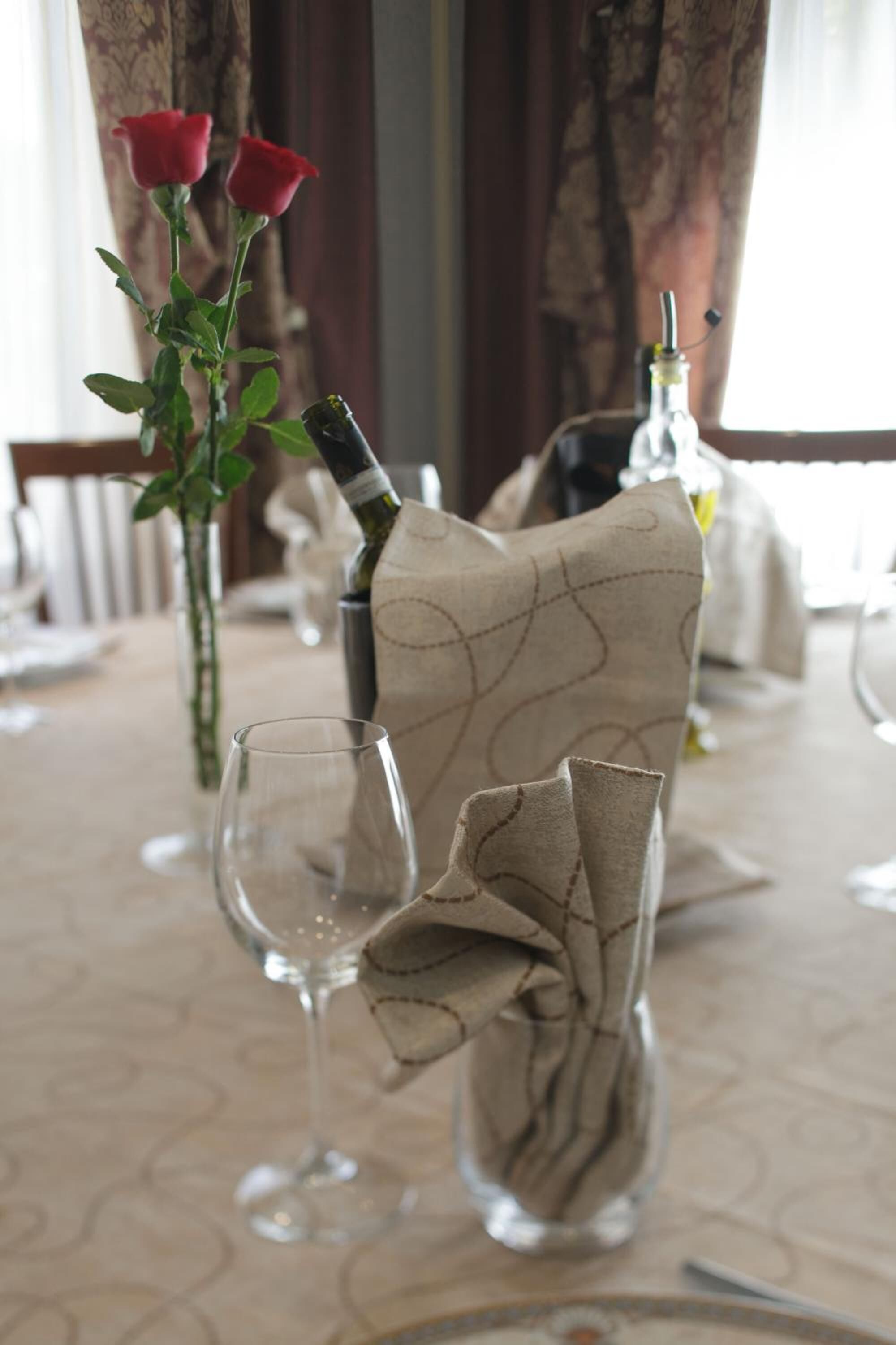 A dining table with a vase of red roses, a bottle of wine, and a cloth-wrapped bottle.