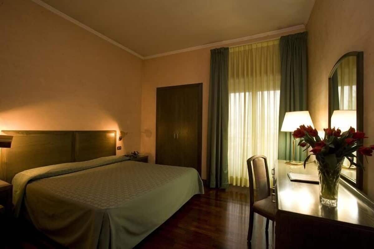 Ein Hotelzimmer mit einem großen Bett, einem Schreibtisch mit Lampe, einer Vase mit Blumen und einem Spiegel.