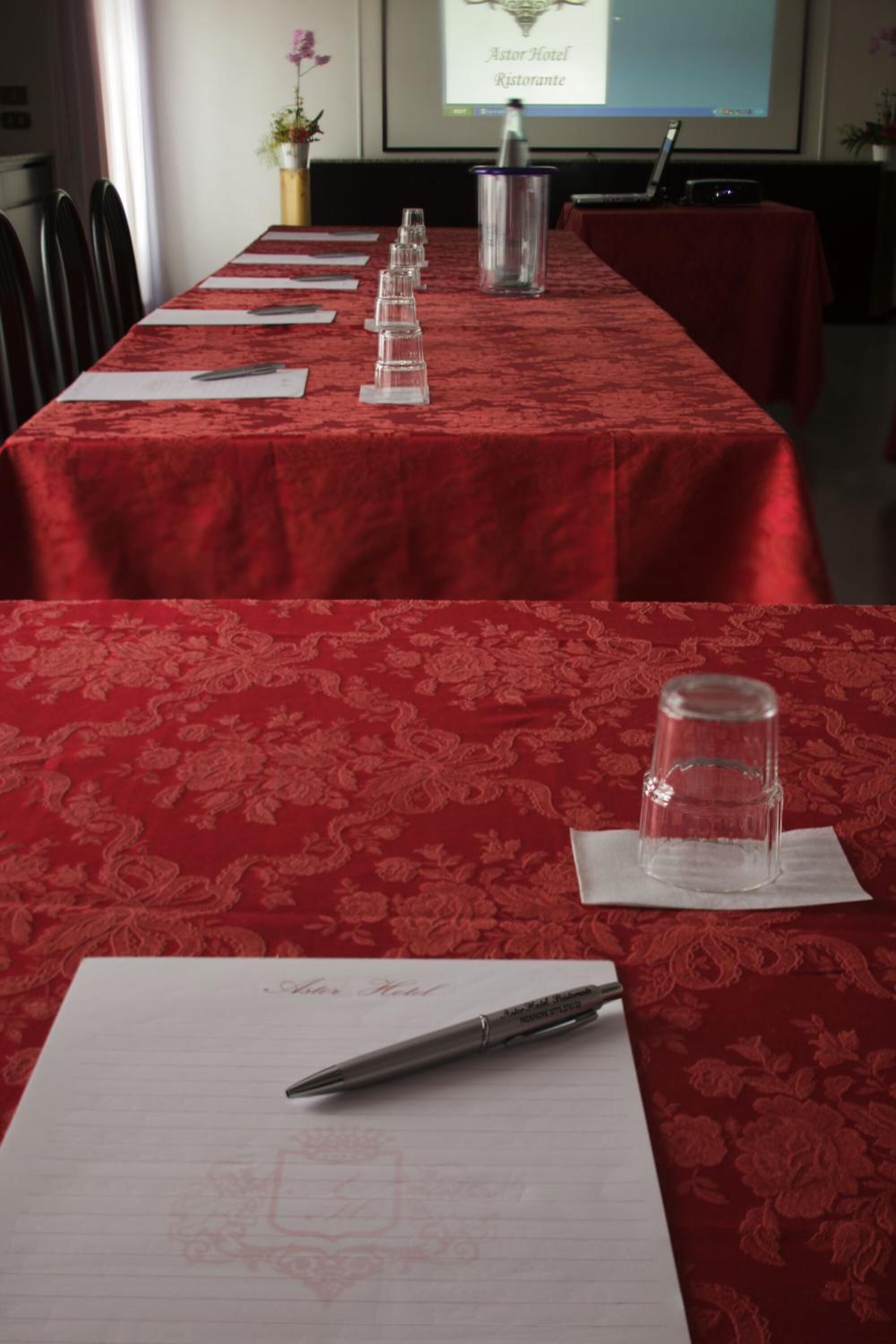 A conference room with a red tablecloth, lined with chairs, a projector screen displaying "Astor Hotel Restaurante", and a laptop on the table.