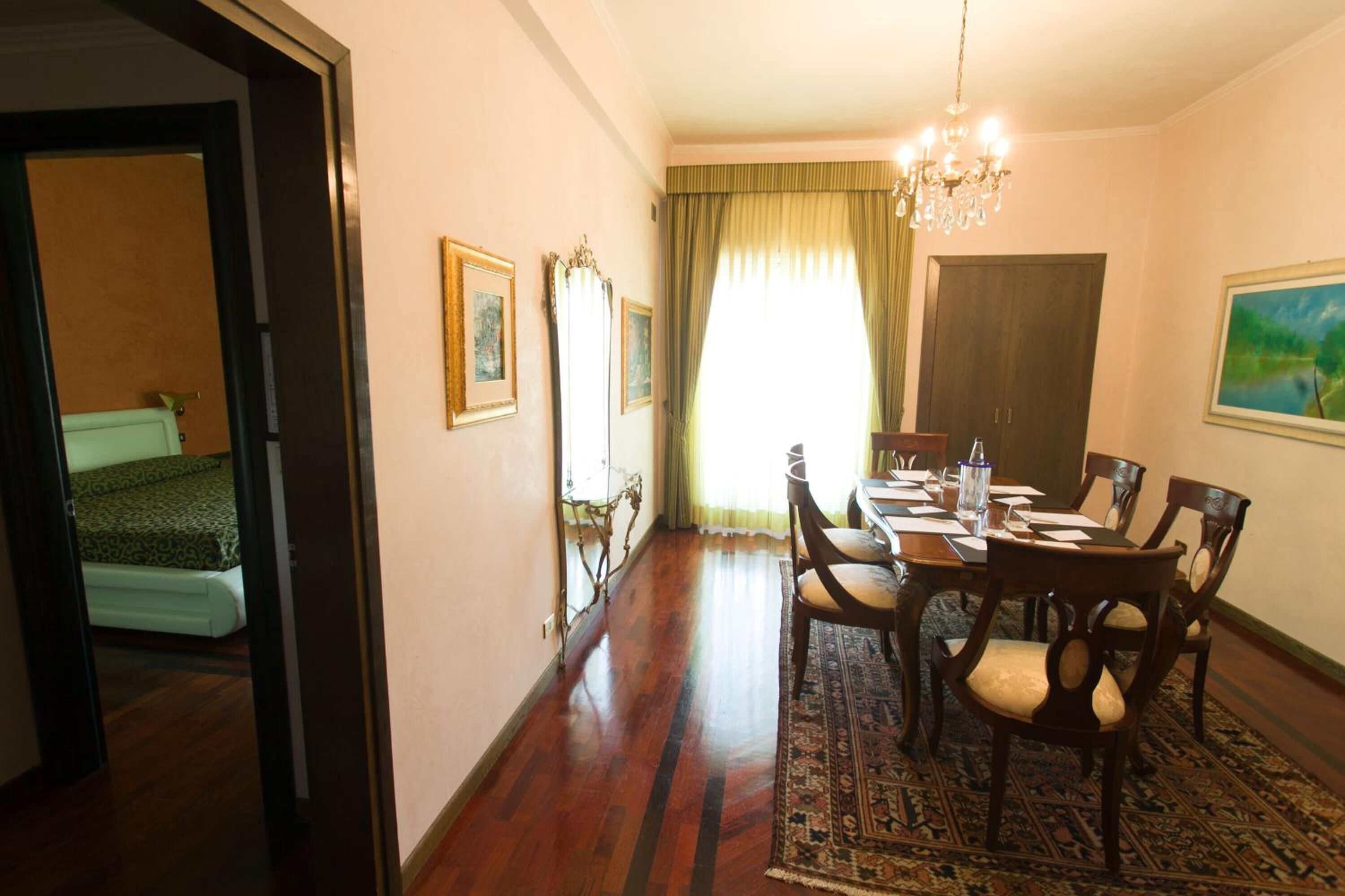 A dining area with a wooden table set for four, surrounded by chairs. A chandelier hangs from the ceiling, and a large window with curtains is visible.