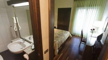Quarto de hotel com piso de madeira, uma cama com colcha florida, uma pequena escrivaninha com cadeira, um espelho e um banheiro com pia e vaso sanitário.