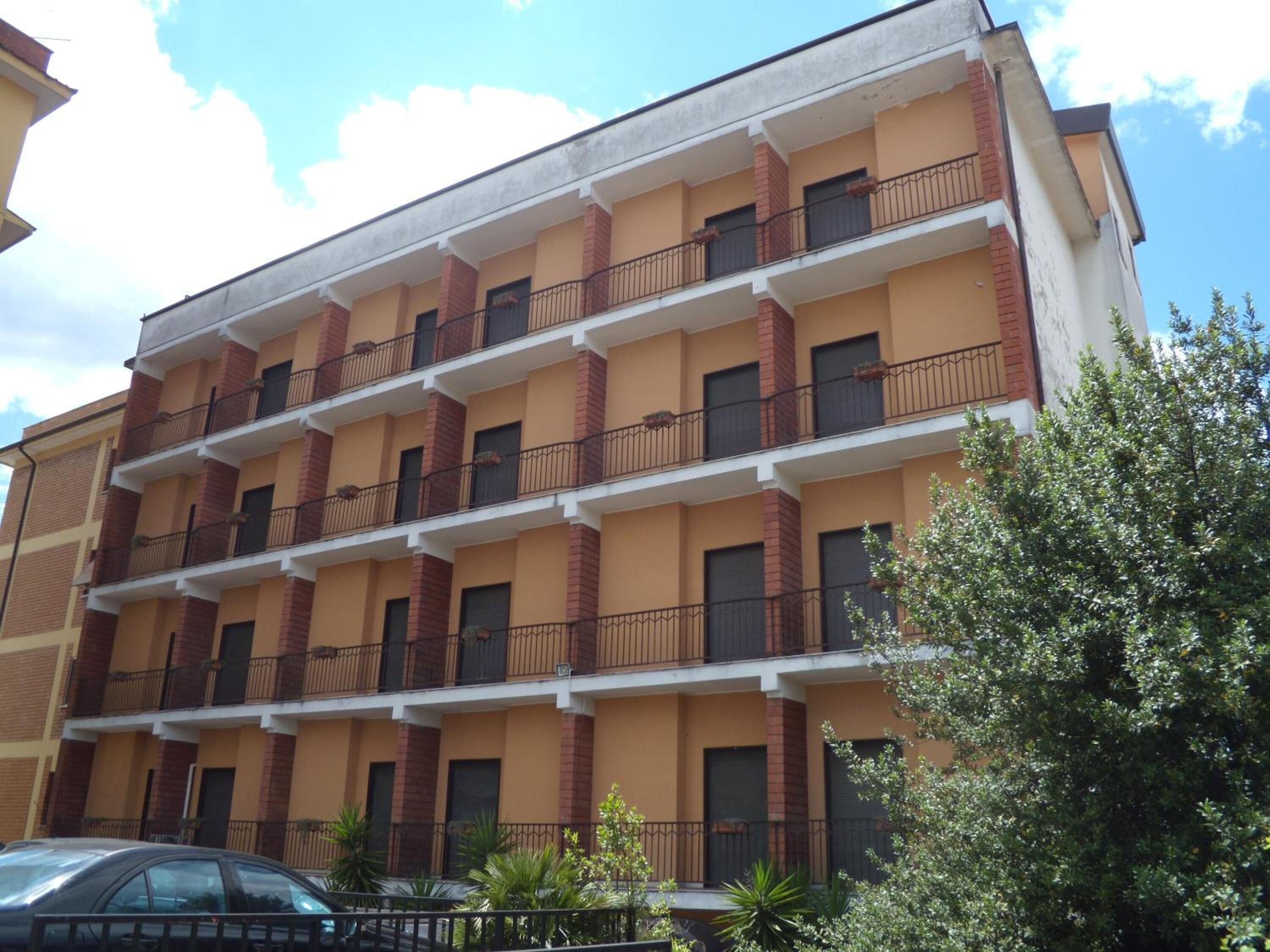 A multi-story building with balconies, a car parked in front, and some greenery.