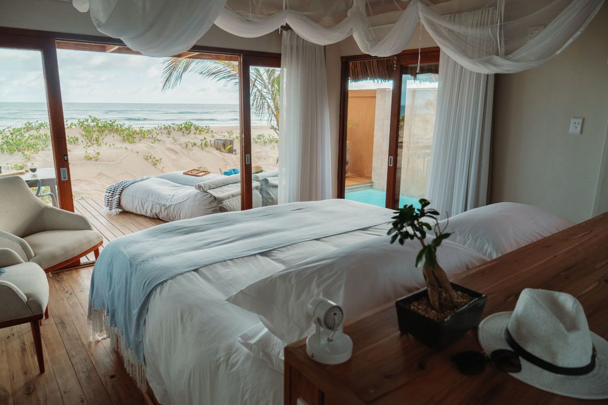 A bedroom with a large bed, a wooden nightstand, a potted plant, and a view of the beach.