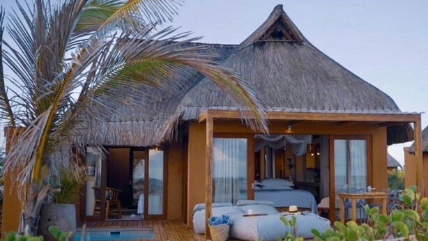 Una cabaña con techo de paja, terraza de madera, piscina y un dormitorio con cama y zona de estar.
