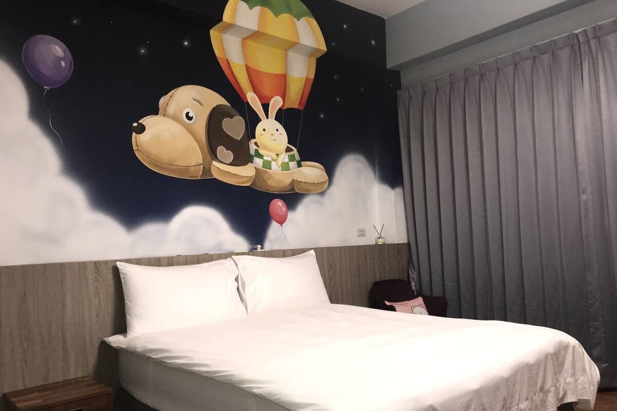 Een slaapkamer met een groot bed, een nachtkastje, een muurschildering van een hond met een luchtballon en een raam met gordijnen.