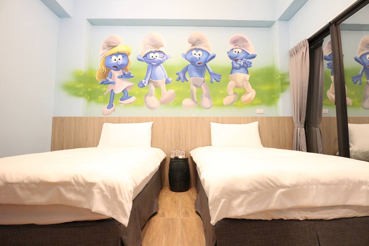 Een hotelkamer met twee bedden, een muurschildering van blauwe figuren aan de wand, en een kleine zwarte prullenbak tussen de bedden.