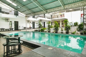 Una piscina interior con una mesa y sillas de vidrio, rodeada de plantas en macetas y una escalera que lleva a un balcón.