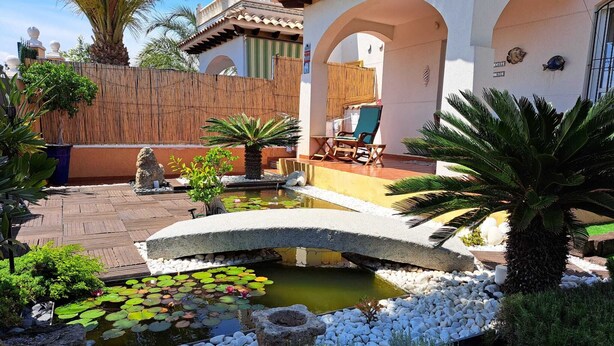 Un patio con un puente de piedra sobre un estanque, rodeado de guijarros blancos y plantas.