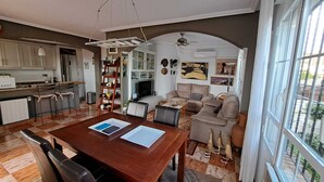 Amplia sala de estar con una mesa de comedor de madera, un sofá y una chimenea. Cuenta con una zona de cocina con barra y un ventanal que ofrece vistas al exterior.
