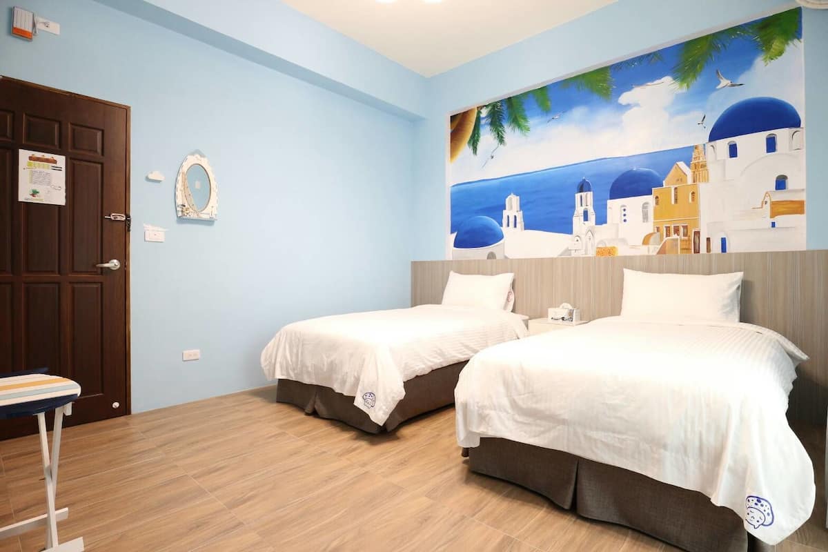 A hotel room with two beds, a wooden floor, a painting of a coastal scene, and a door with a sign.