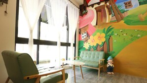 Ein Zimmer mit einem grünen Sofa, einem Holzstuhl, einem kleinen Tisch und einem Wandgemälde, das eine Landschaft mit Blumen und einem Haus zeigt.