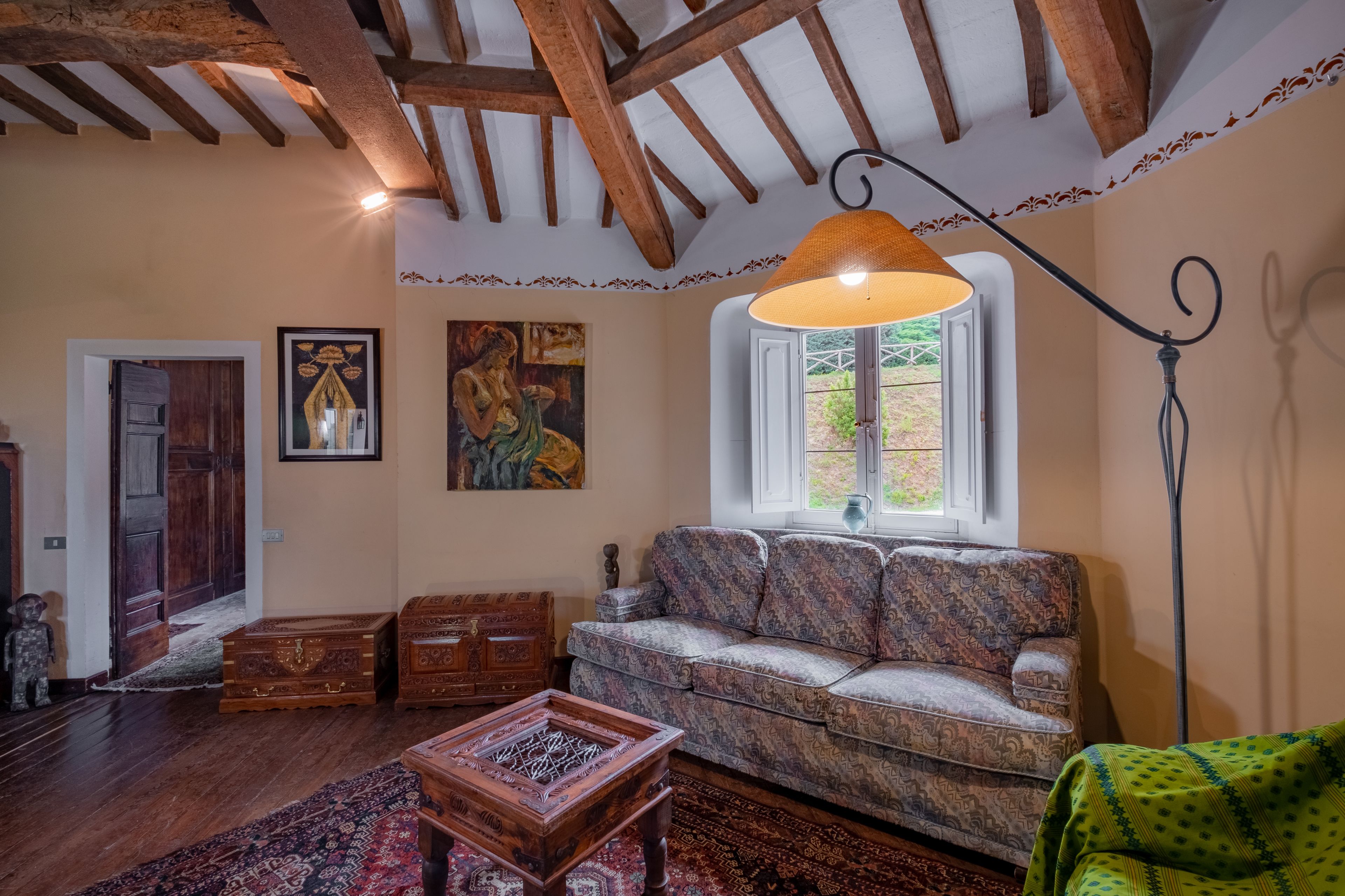 Un salon chaleureux avec un canapé à motifs, des meubles en bois, une grande fenêtre offrant une belle vue et un plafond avec des poutres apparentes.