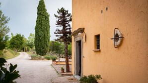 A yellow building with a gravel driveway leading to a door, surrounded by potted plants and a tall tree. - Agriturismo Le Cerque (Collazzone)