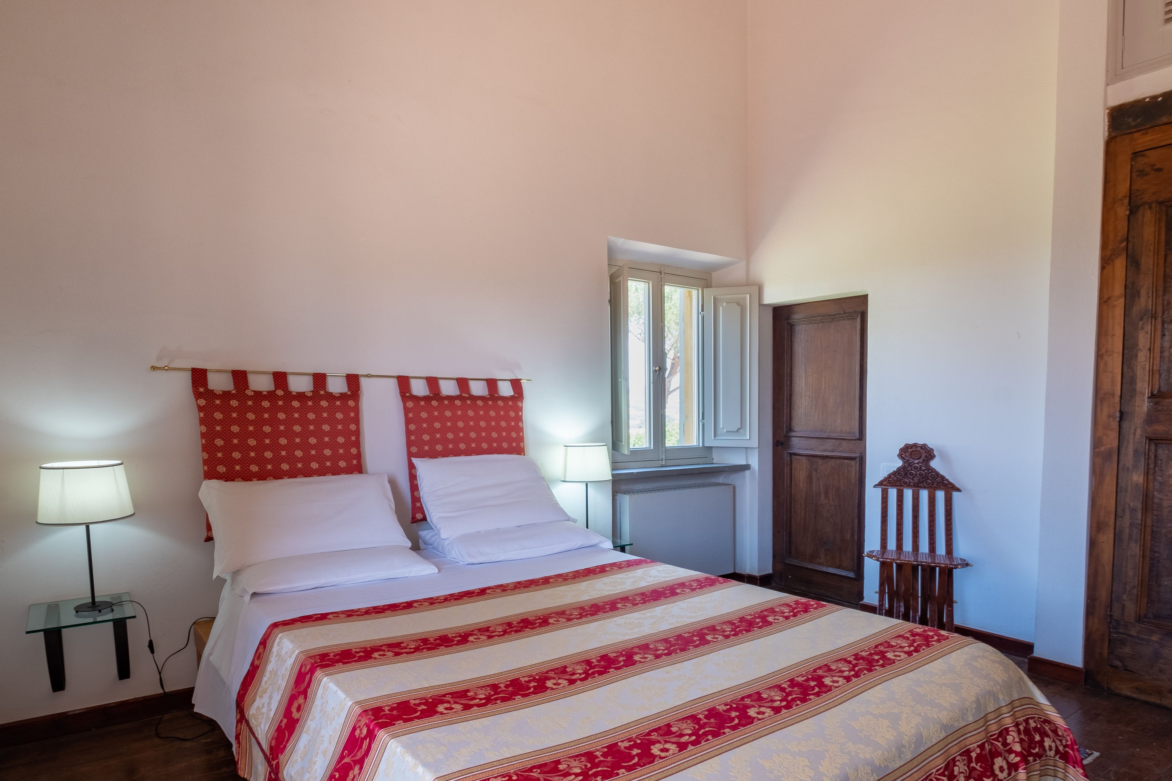 Un letto ben rifatto con copriletto rosso e bianco a motivi decorativi, una lampada da comodino e un comodino in legno.