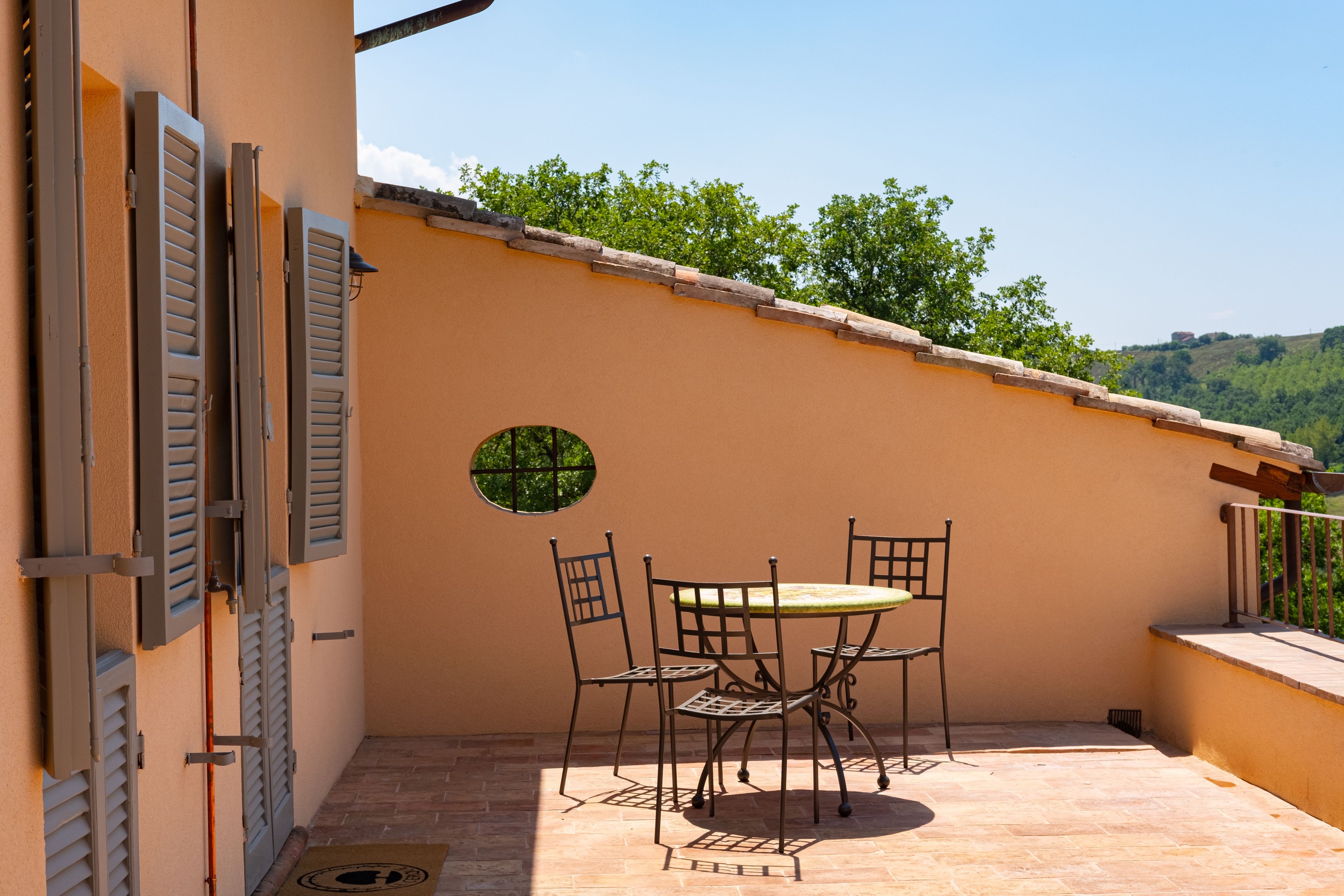 A patio with a table and chairs, a round window, and a view of greenery and hills.