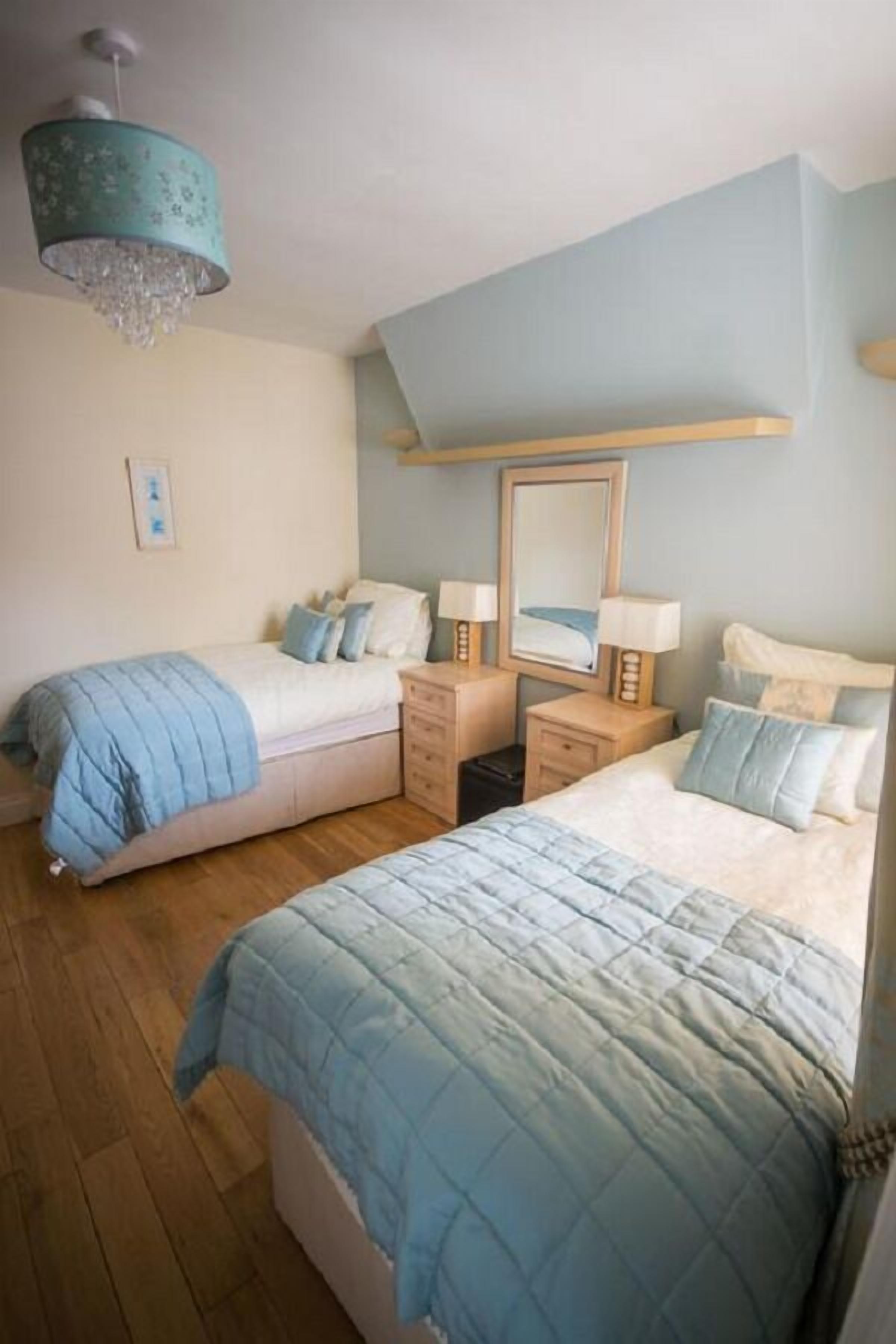 A bedroom with two beds, wooden flooring, a ceiling light fixture, and a mirror above the beds.