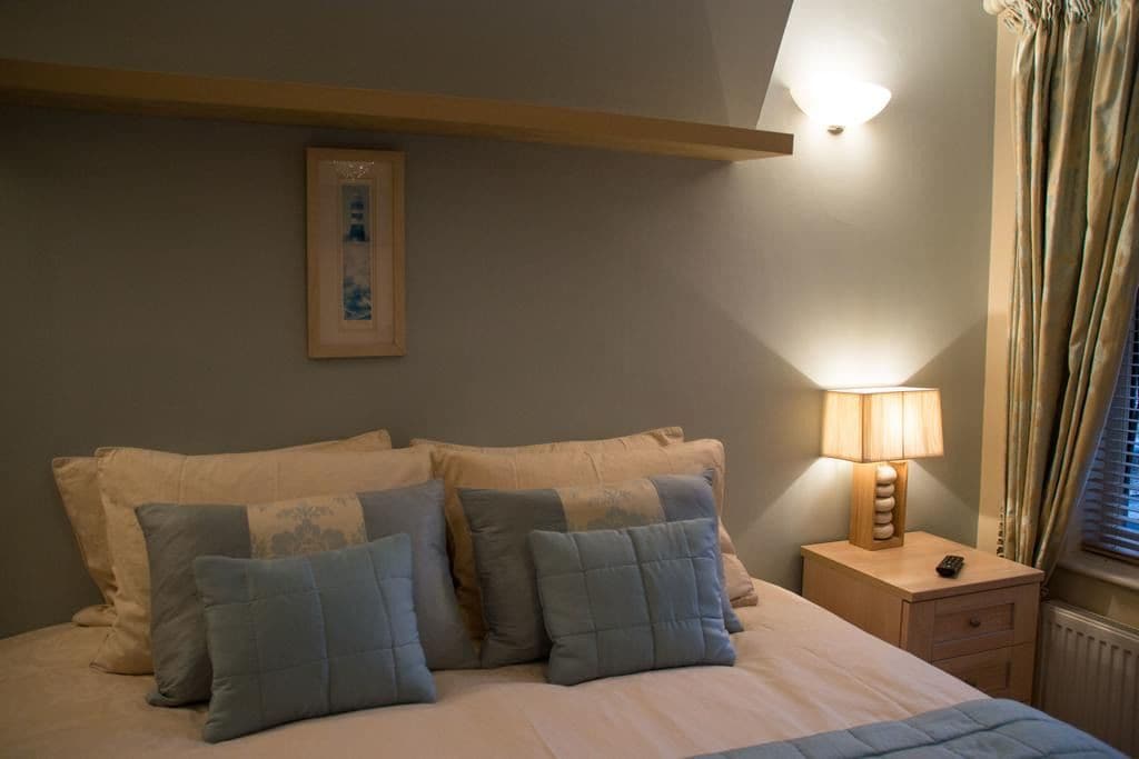 A bedroom with a bed, pillows, a bedside table with a lamp, a framed picture on the wall, and a window with curtains.