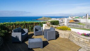 Een terras met rieten meubilair, een stenen muur en uitzicht op zee en de stad.