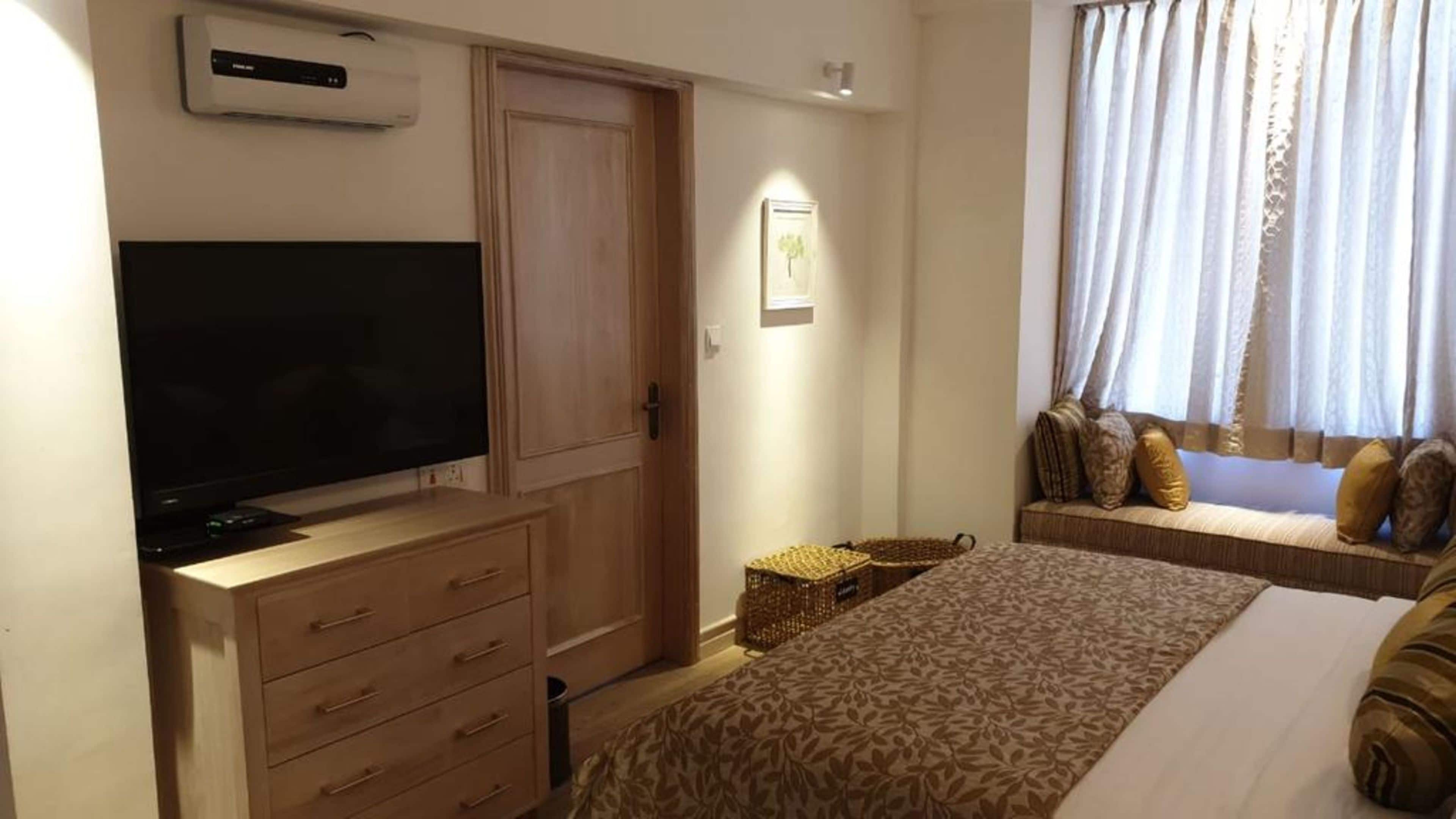 Une chambre avec une télévision, une armoire en bois, une table de chevet et une fenêtre avec des rideaux.