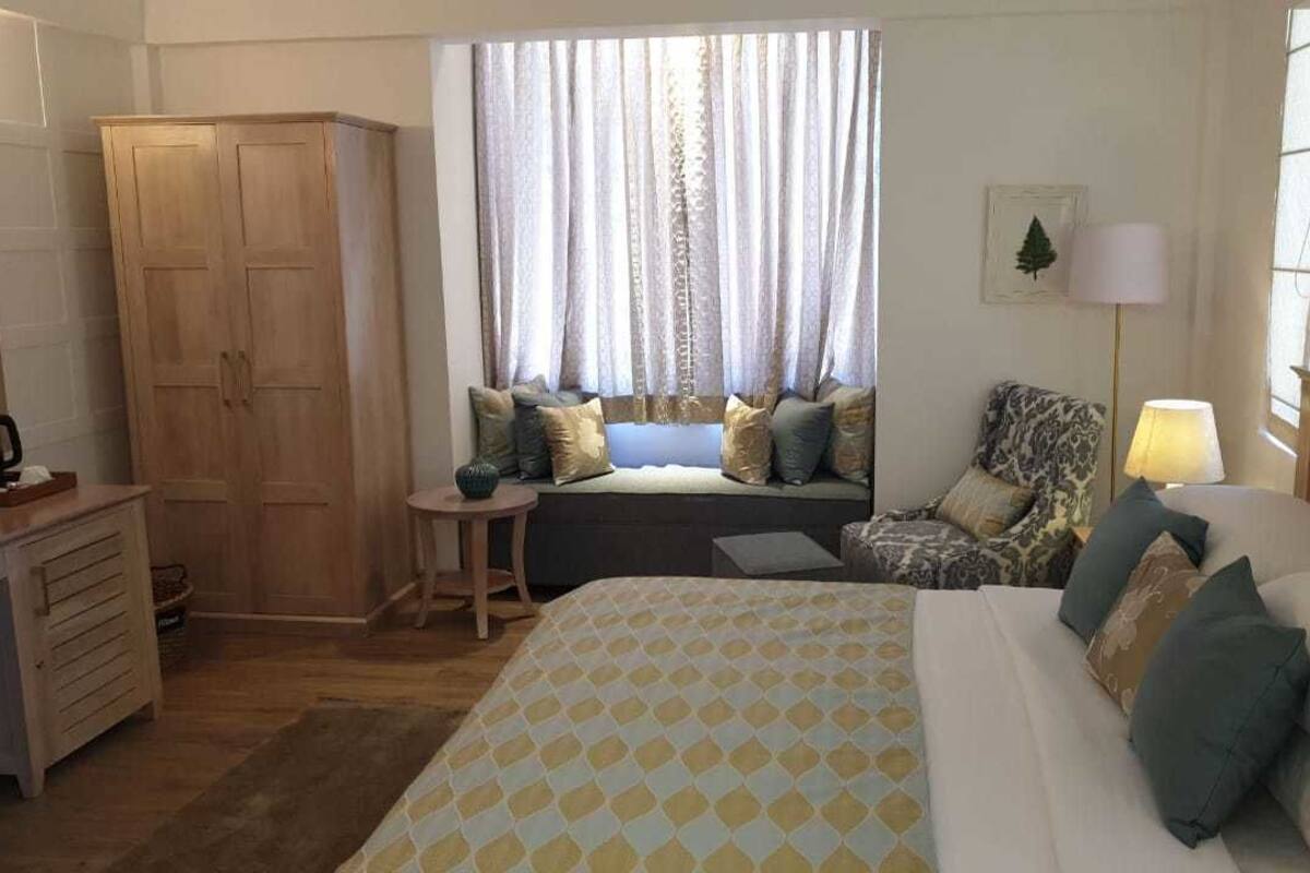 Une chambre d’hôtel comprenant un lit, un coin salon avec des coussins, une armoire, un bureau avec une lampe et une fenêtre avec des rideaux.