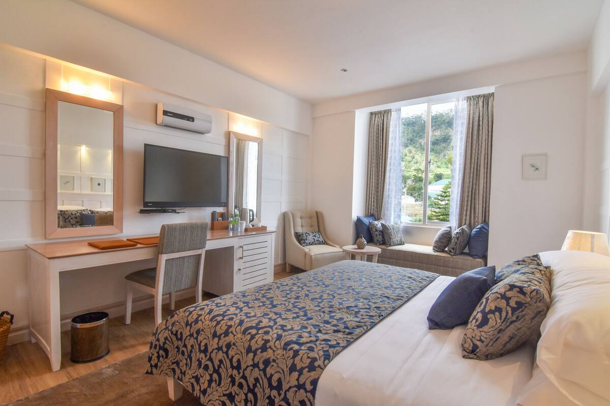 Une chambre d’hôtel avec un lit, un bureau avec une télévision, une chaise, une fenêtre avec des rideaux et une vue sur la verdure.