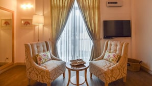 Une chambre d’hôtel avec deux fauteuils rembourrés, une petite table ronde, un téléviseur fixé au mur et une grande fenêtre ornée de rideaux légers.