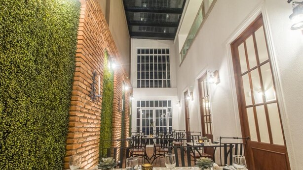 Un espace repas avec des tables préparées pour deux personnes, un mur en briques agrémenté de plantes et de grandes fenêtres.
