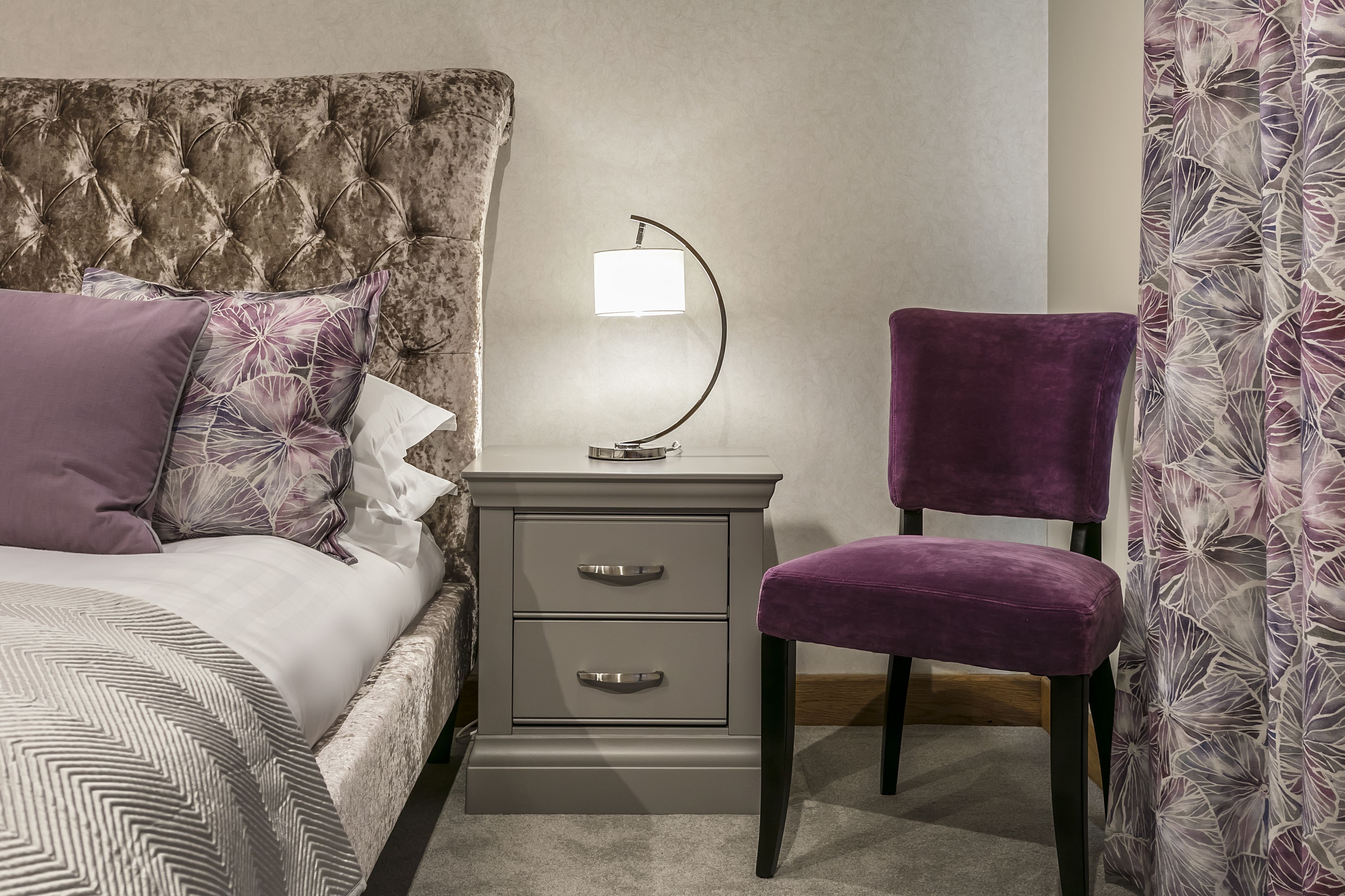 A bedroom with a tufted headboard, a bedside table with drawers, a lamp, a purple chair, and a floral-patterned curtain.