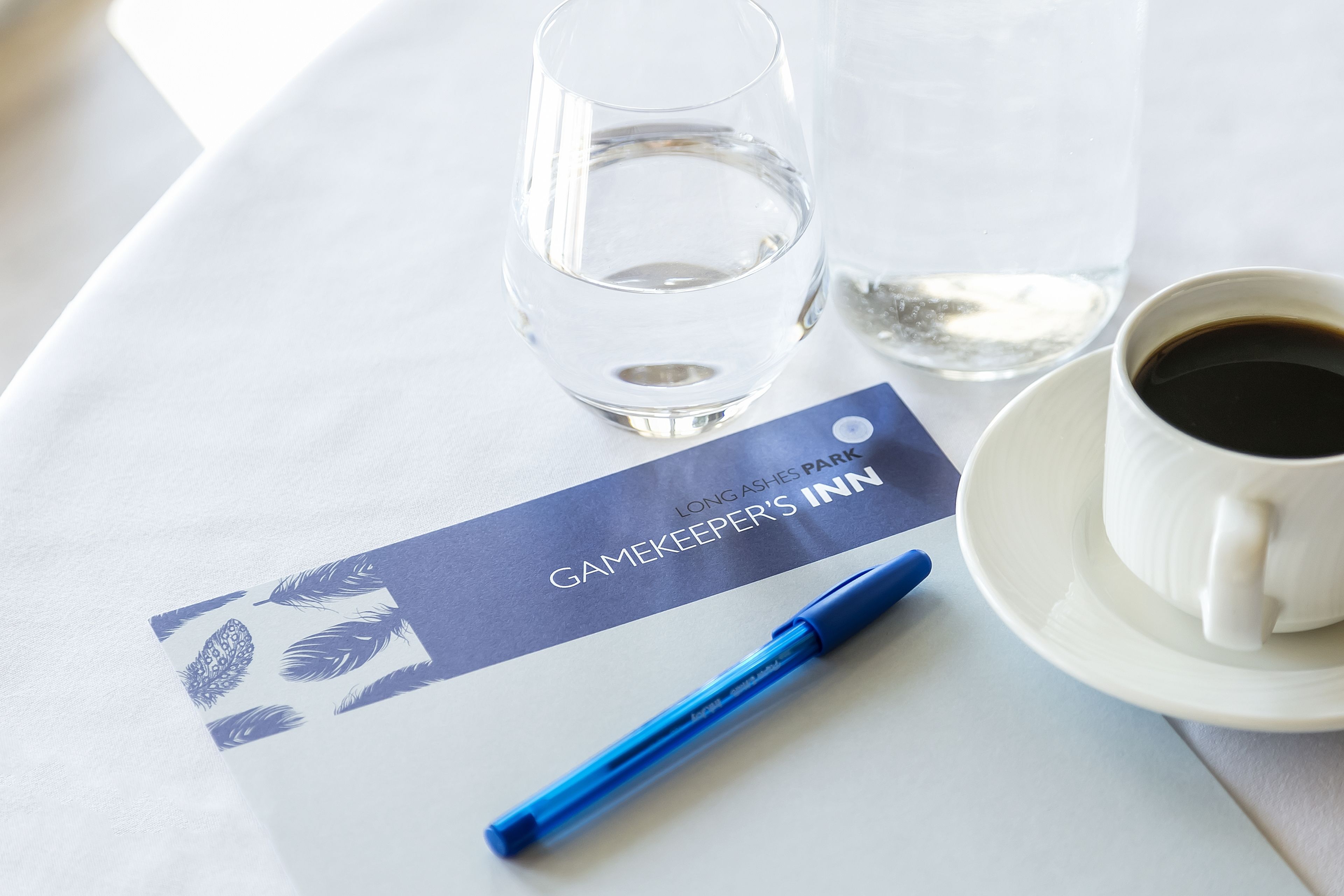 Une table avec un verre d’eau, une tasse de café, un stylo et une carte portant l’inscription « Auberge du gardien de chasse ».