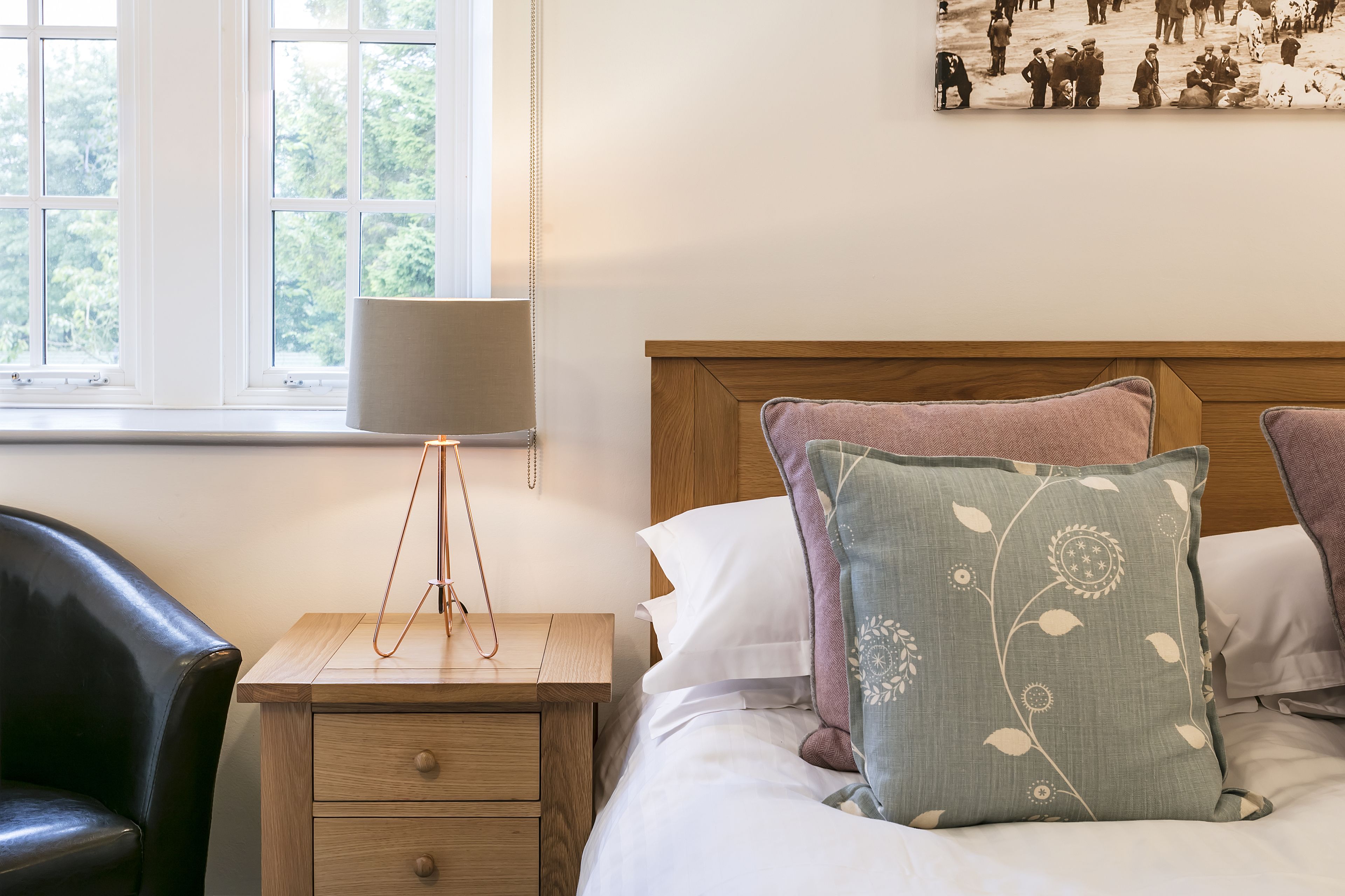 Une chambre avec une table de chevet en bois, une lampe, un lit agrémenté d’oreillers à motifs floraux, un fauteuil en cuir noir et une fenêtre donnant sur des arbres.