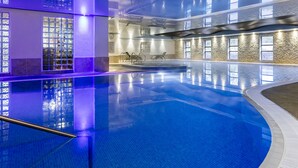 Una piscina interior con agua azul cristalina, suelo de baldosas y un diseño moderno con pared de piedra y ventanas.