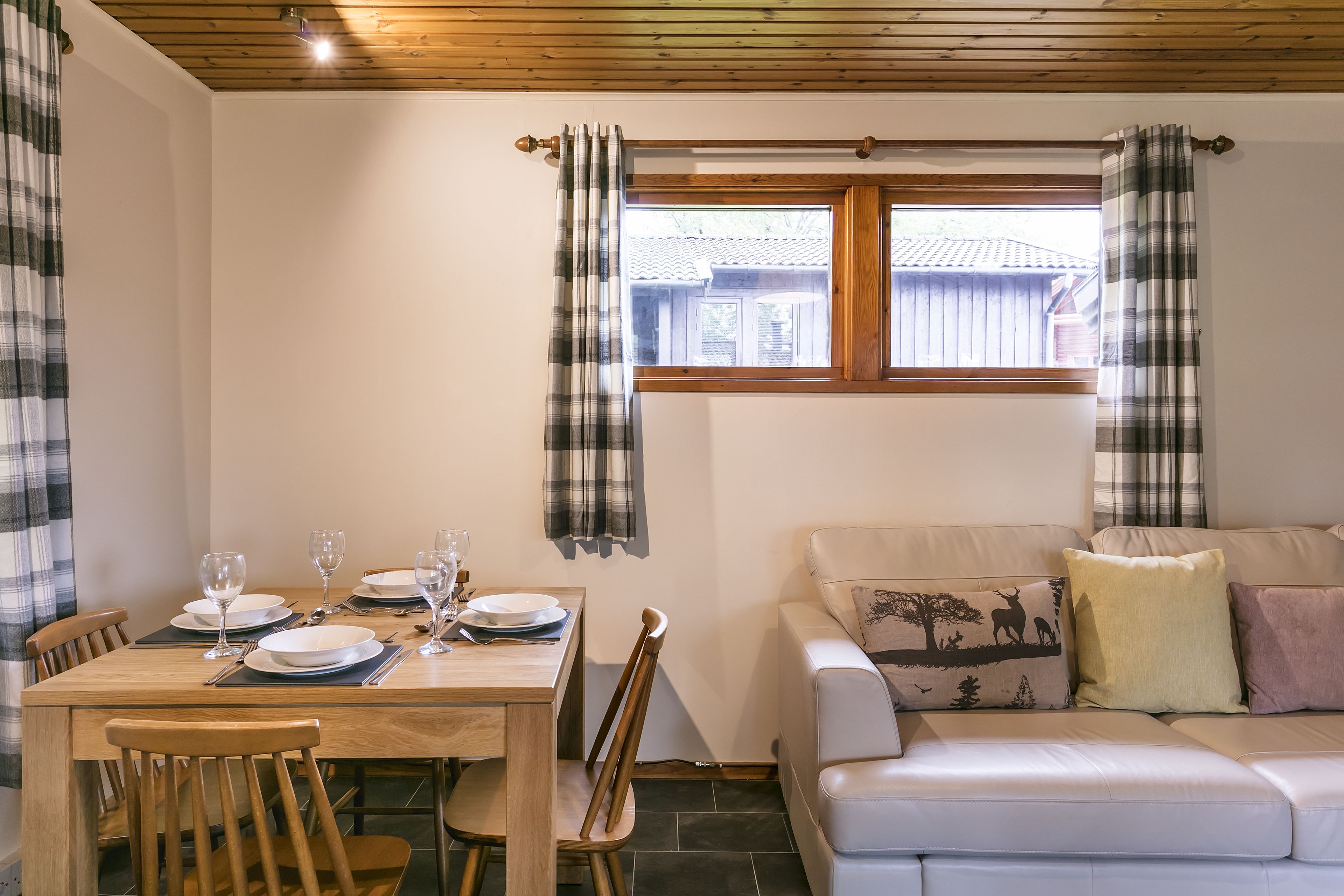 A dining area with a wooden table set for four, a sofa with decorative pillows, and a window with plaid curtains.