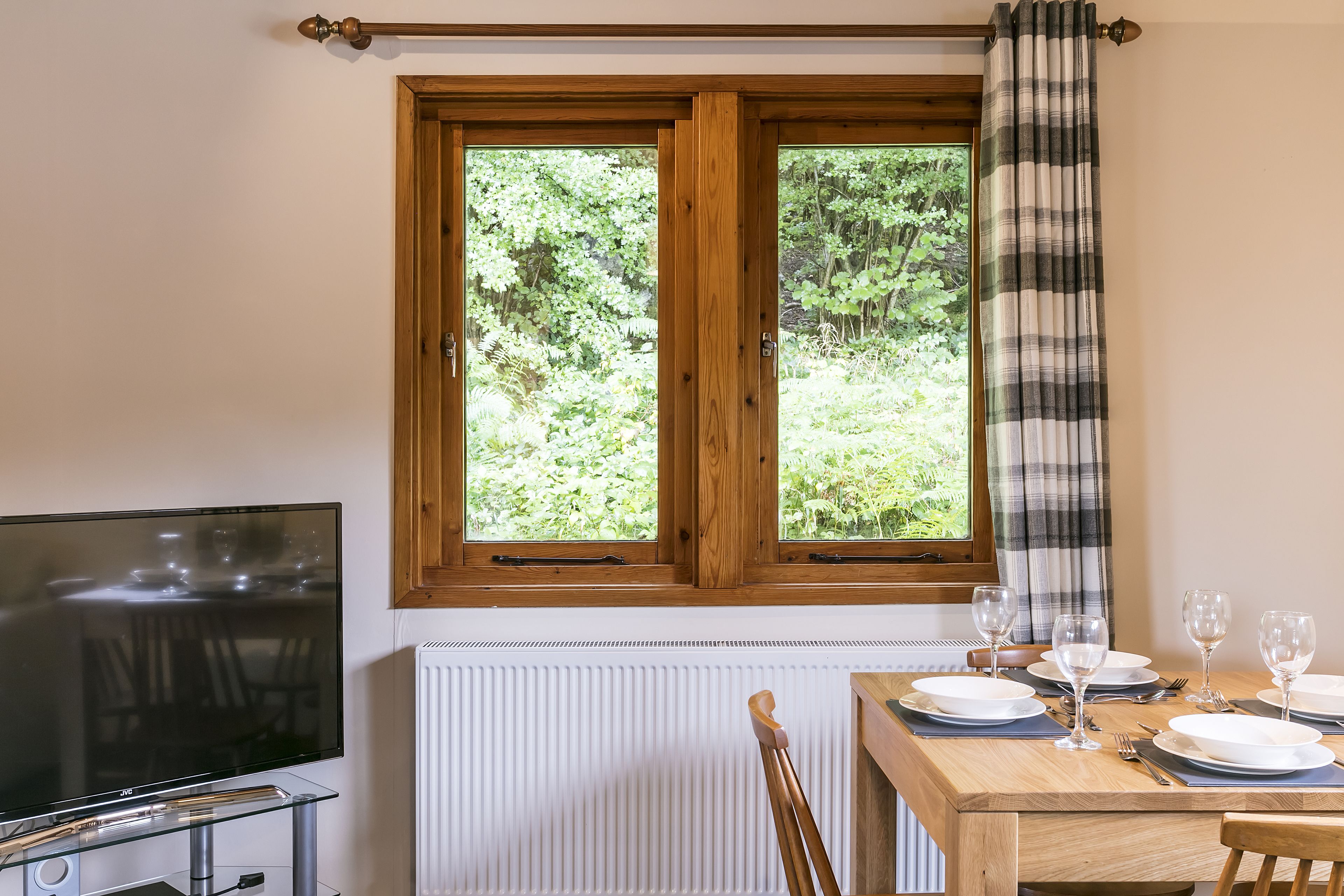 A dining area with a wooden table set for four, a television on a glass stand, and a window with a plaid curtain.