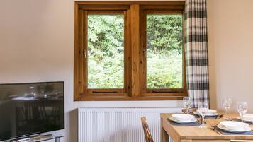 A dining area with a wooden table set for four, a television on a glass stand, and a window with a plaid curtain.