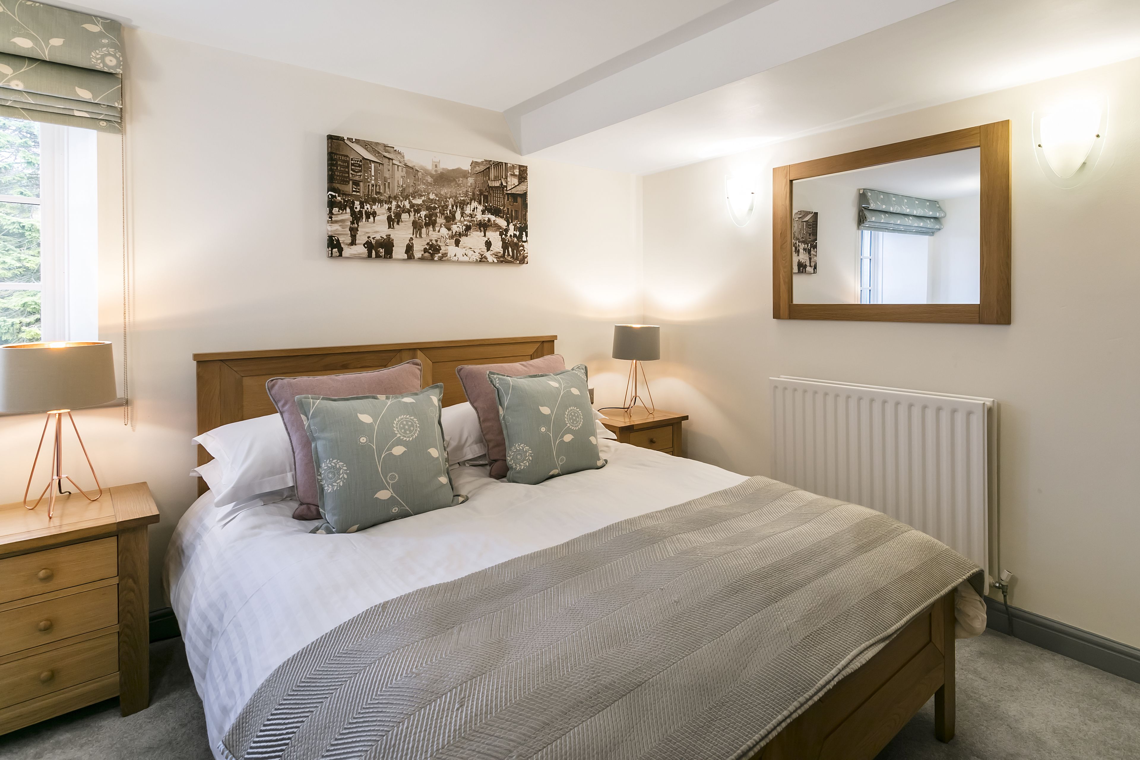 A neatly arranged bedroom with a large bed, a bedside table with a lamp, a wall-mounted mirror, and a framed picture above the bed.