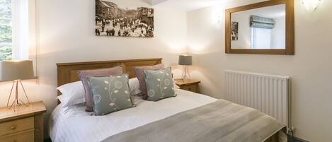 A neatly arranged bedroom with a large bed, a bedside table with a lamp, a wall-mounted mirror, and a framed picture above the bed.