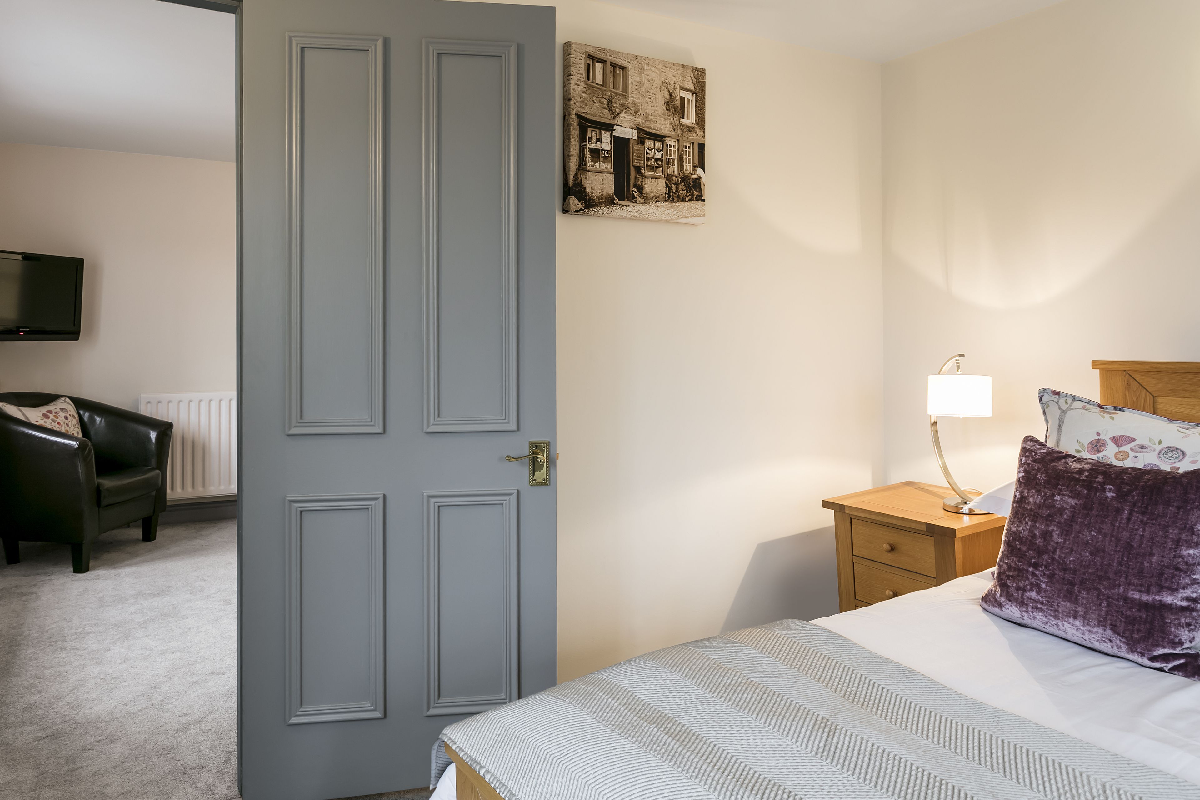 A bedroom with a bed, a nightstand, a lamp, a TV, and a door.