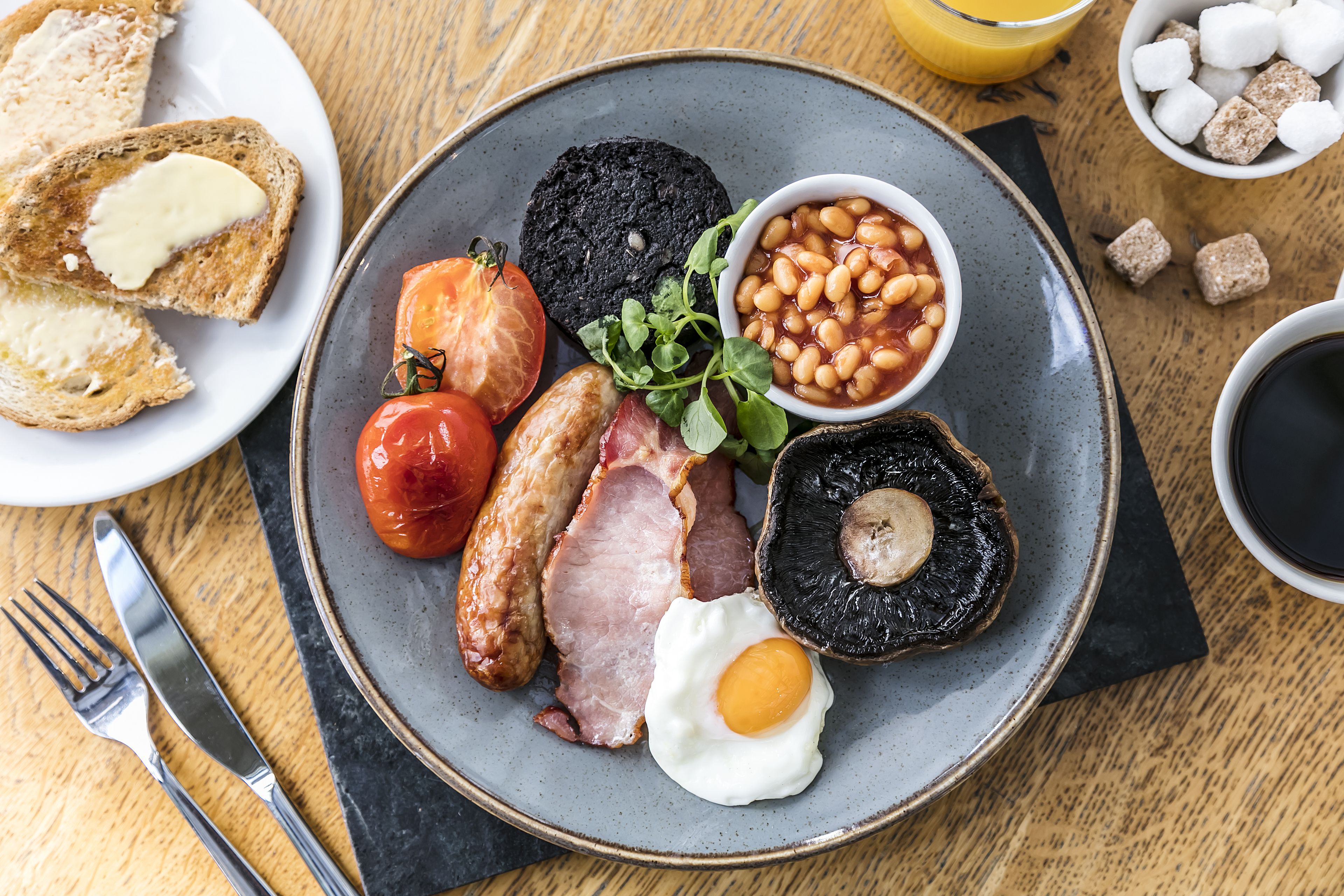 A breakfast plate with toast, eggs, bacon, tomatoes, mushrooms, beans, and a cup of coffee.