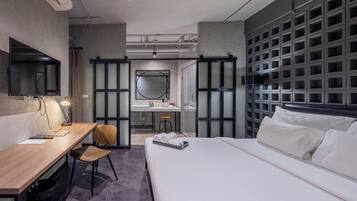 A modern hotel room with a wooden desk, a chair, a bed with white linens, a large mirror, a bathroom with a sink and a shower, and a wall with a grid of small windows.