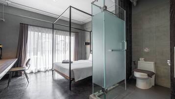 A modern hotel room with a glass partitioned shower, a bed, a desk with a chair, and a view of the city through large windows.