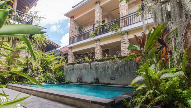 A pool area with a stone wall and tropical plants, a balcony with a railing, and a building with columns. - OYO 401 The Frog Homestay Sanur (Denpasar)