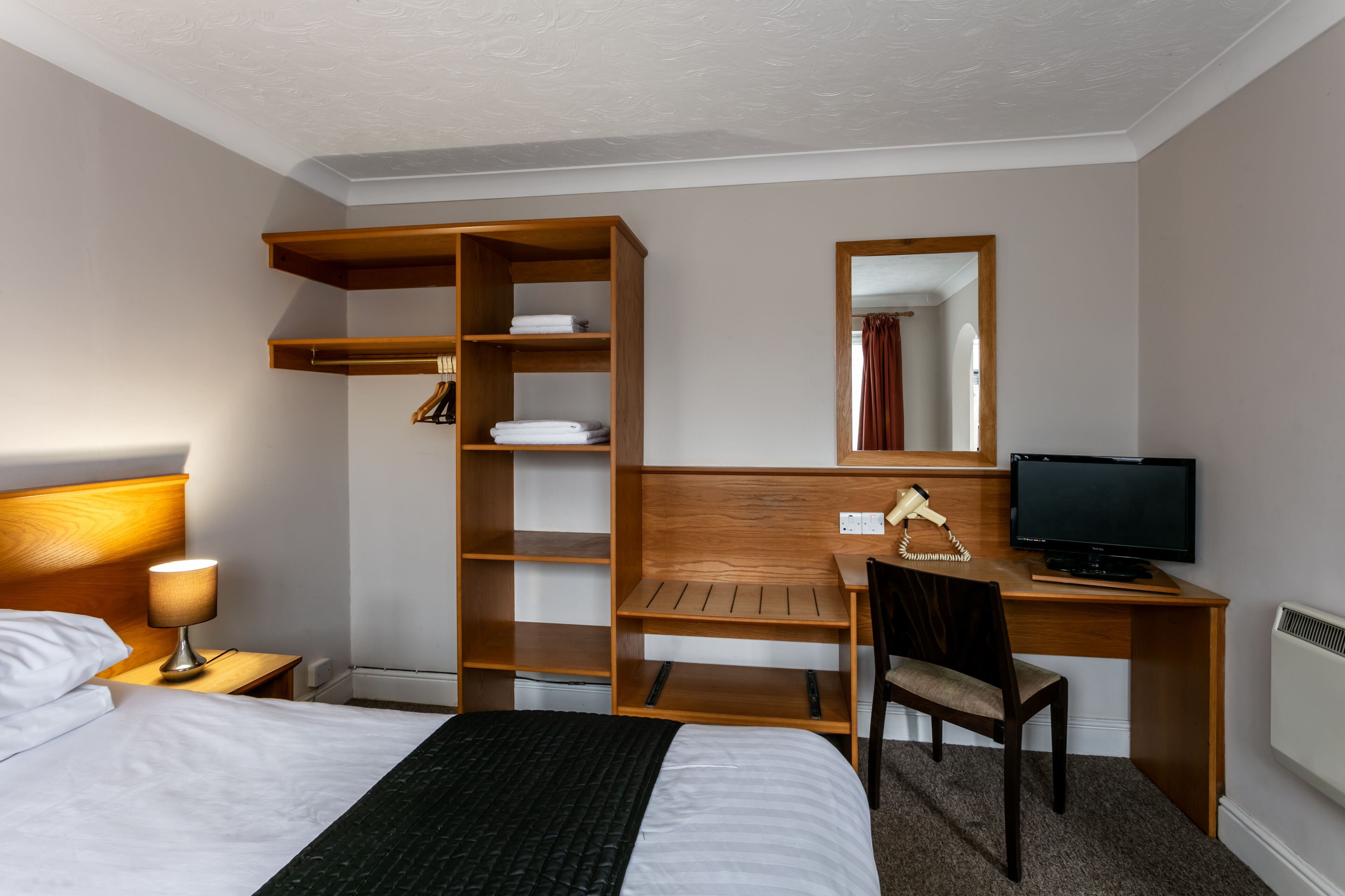 A hotel room with a bed, a desk with a computer, a chair, a TV, a lamp, and built-in shelves.