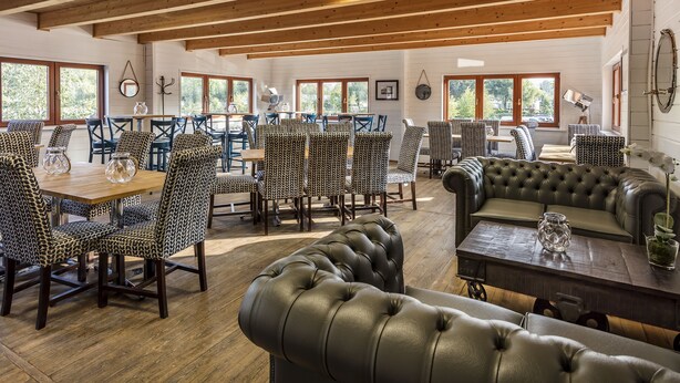 A dining area with wooden tables and chairs, a leather sofa, and large windows with a view of greenery. - Roydon Marina Village Hotel (Harlow)
