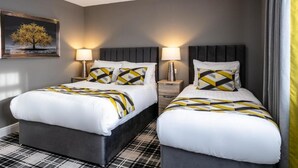 A hotel room with two beds, each with a black headboard, white bedding, and geometric-patterned pillows. There are bedside tables with lamps, a painting on the wall, and a checkered floor. - Roydon Marina Village Hotel (Harlow)