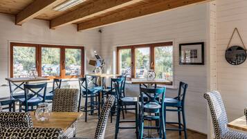 A dining area with wooden tables and chairs, large windows with views of the outdoors, and a wooden ceiling.