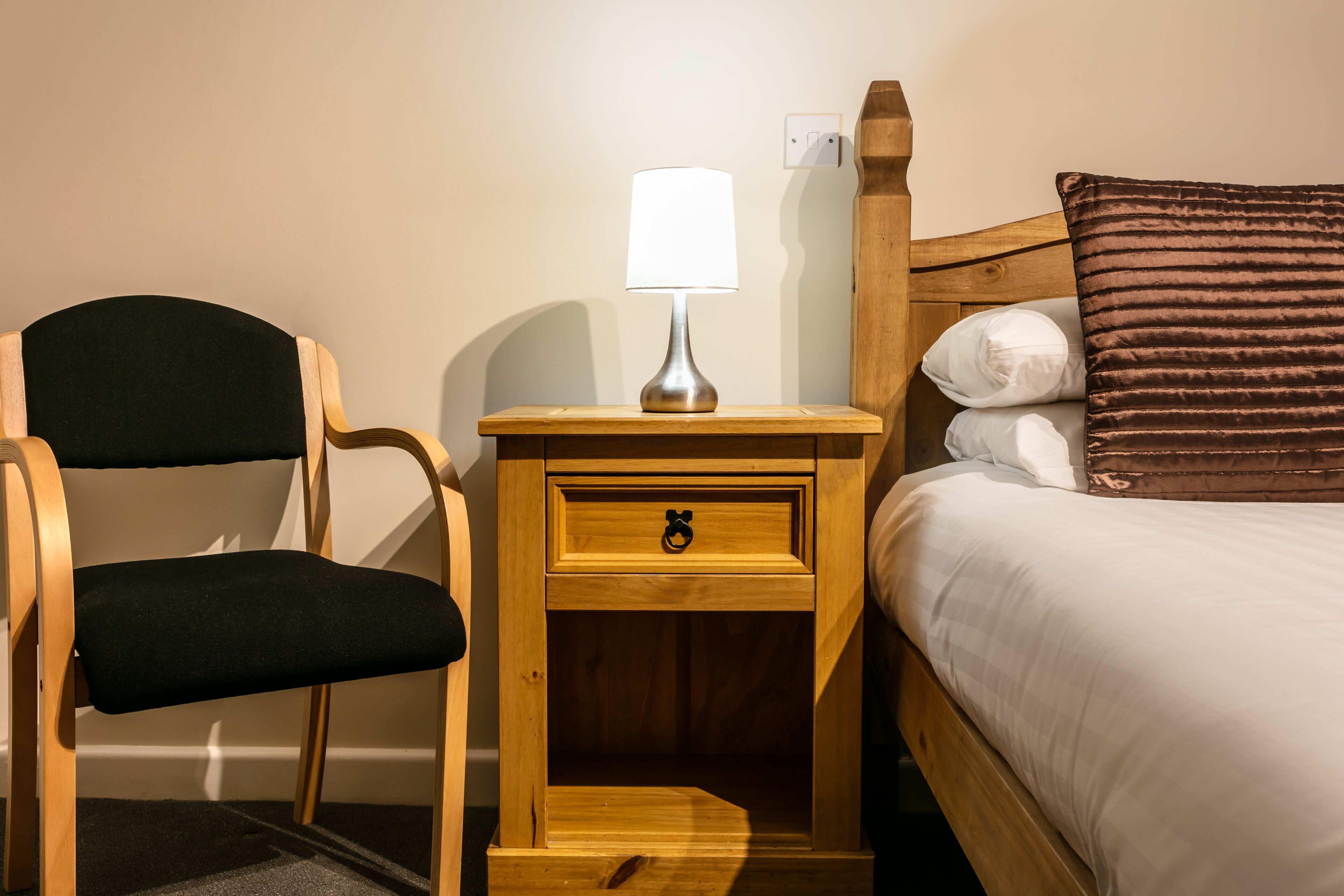 A wooden chair with a black cushion, a bedside table with a lamp, a drawer, and a bed with a brown pillow.