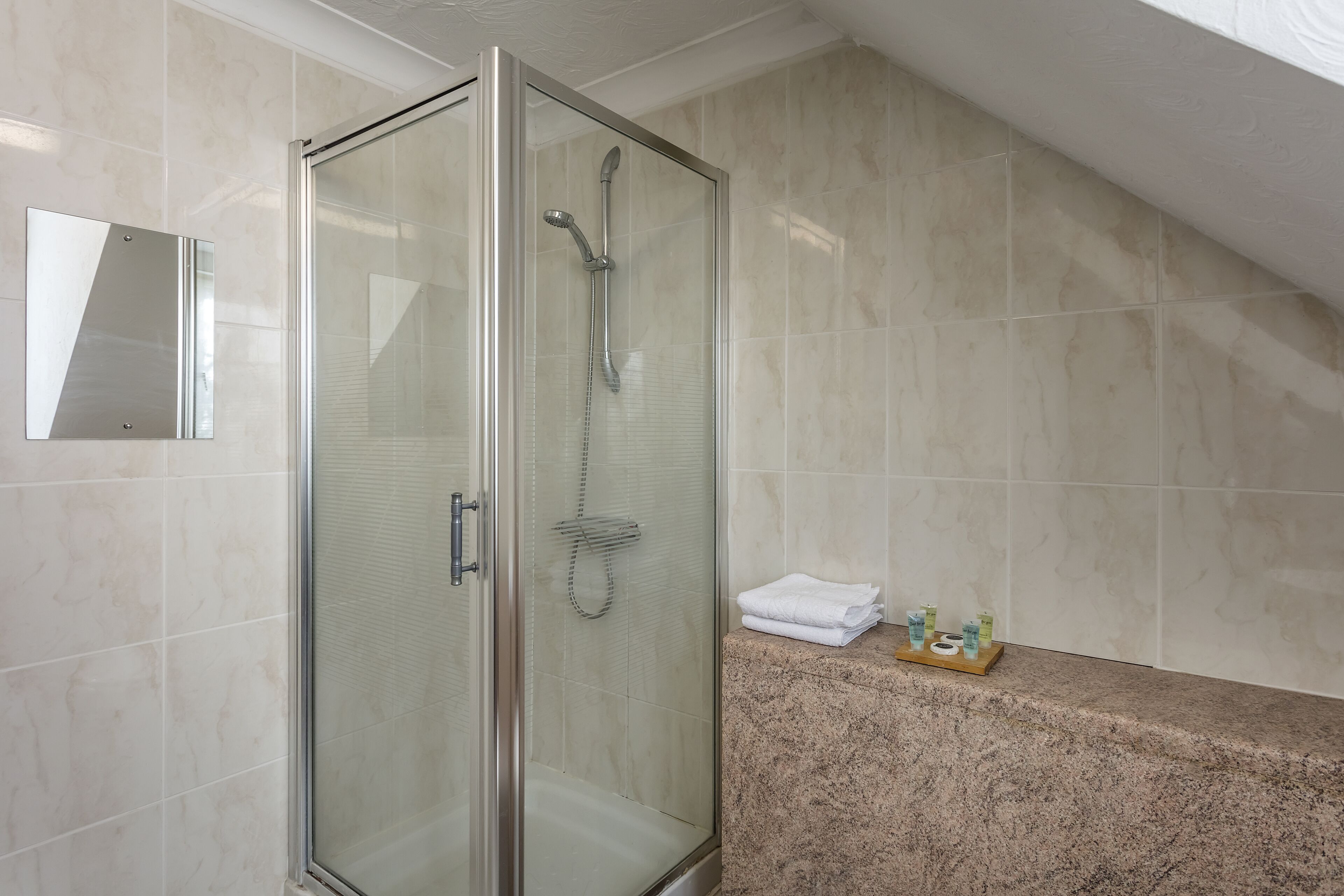 Een moderne douchecabine met een glazen deur, een wand betegeld met marmer en een granieten aanrecht met toiletartikelen.